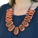 Este colar maxi destaca-se pelo contraste vibrante entre o tom terracota/laranja suave e a riqueza da madeira natural em tom castanho escuro.